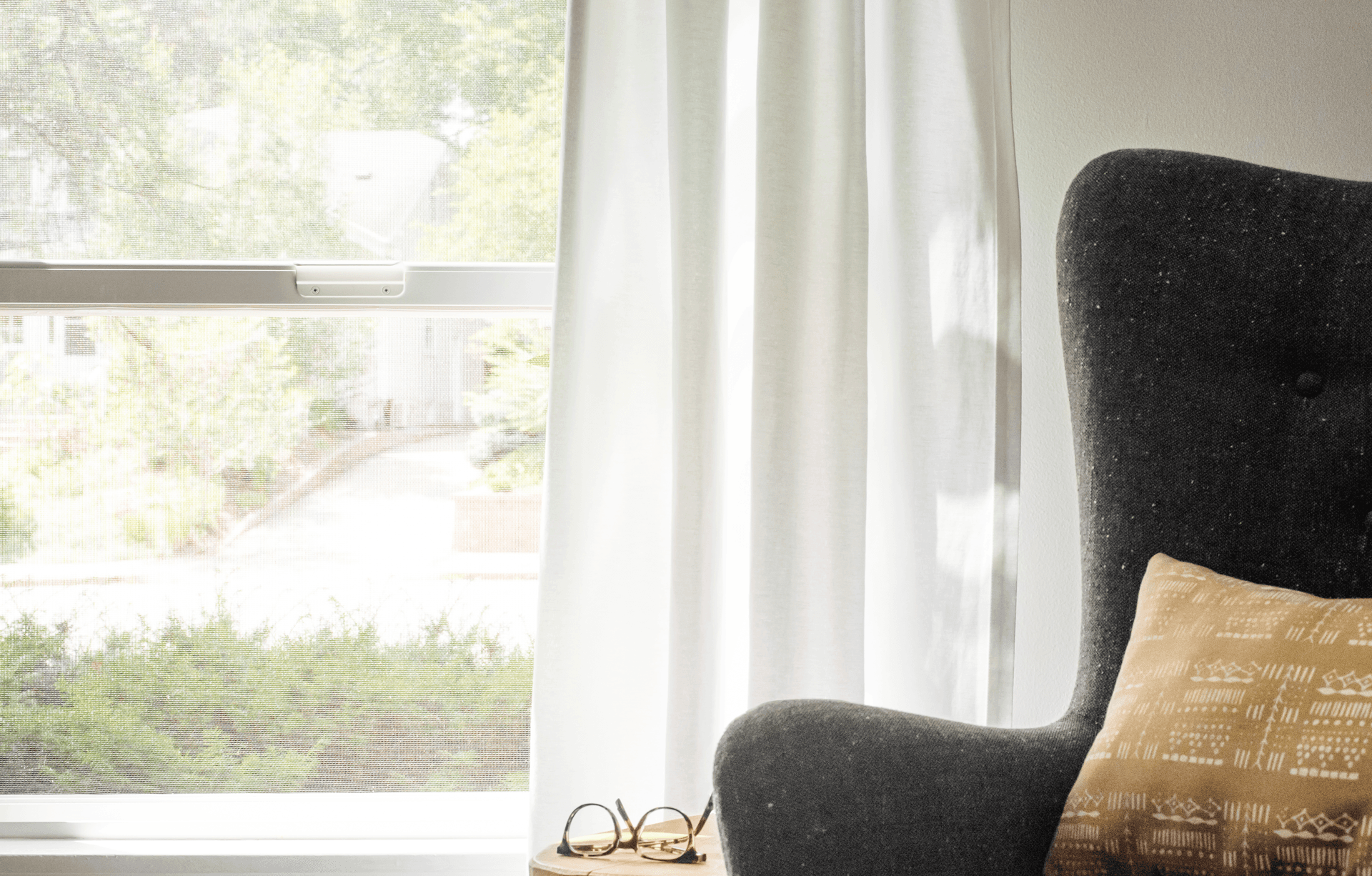 A close up of a window letting light into the room with a chair and a pair of glasses on an end table.