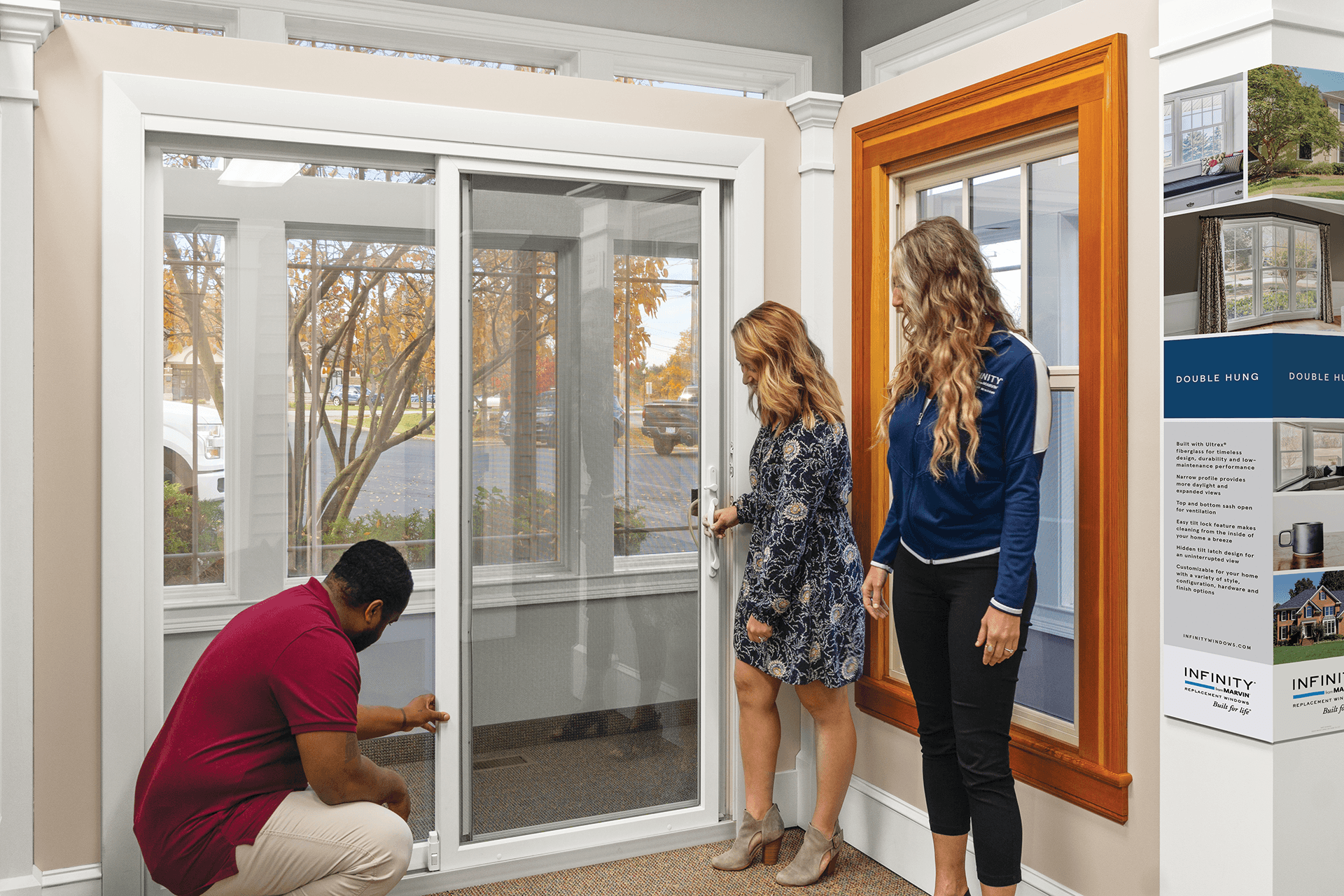 A blonde woman tests a white Infinity sliding patio door with a man bending down with a salesperson watching.