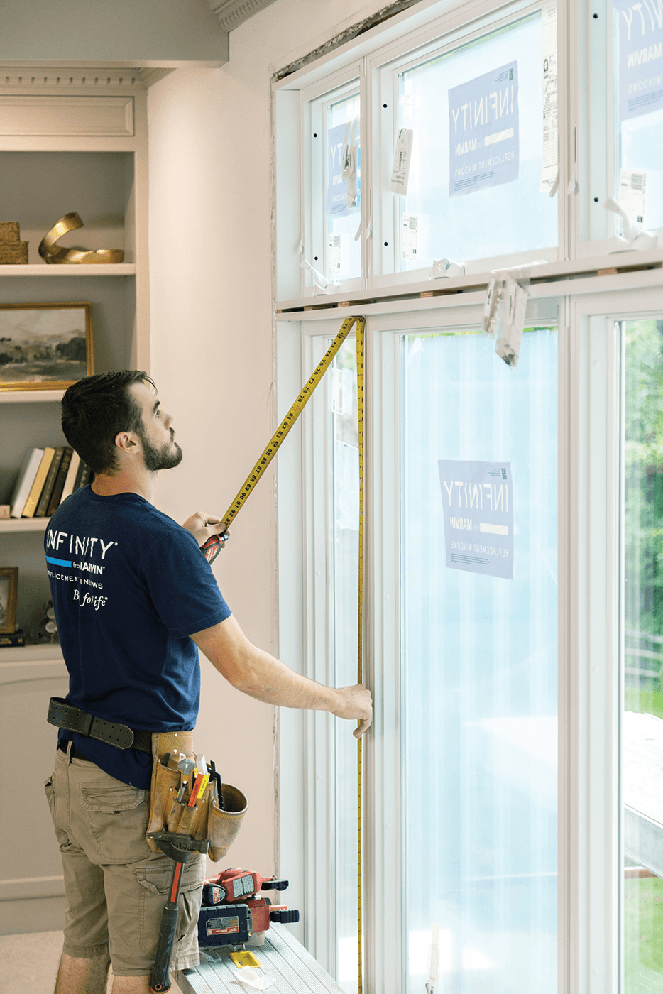 A window installer in a blue shirt uses a tape measure to check the height of an installed window.