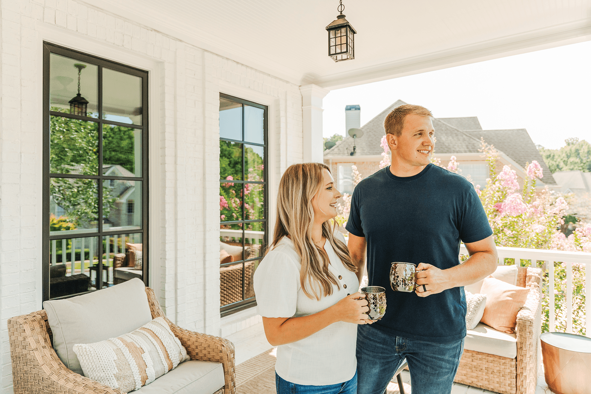A blonde woman stands next to a blond man on a porch holding a copper mug in front of black Infinity casement windows.