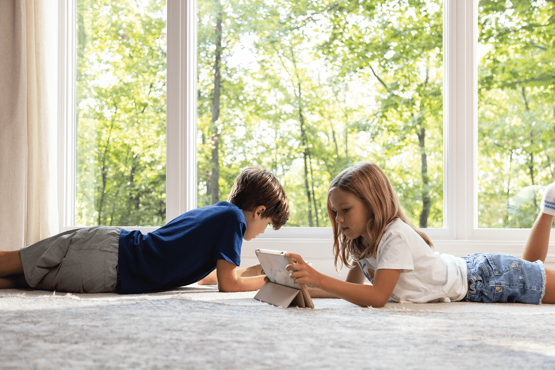 A young boy and young girl lay on the floor inside a home in front of white Infinity picture windows.